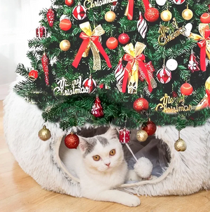 Cat peeking out from a cat house underneath a decorated Christmas tree.