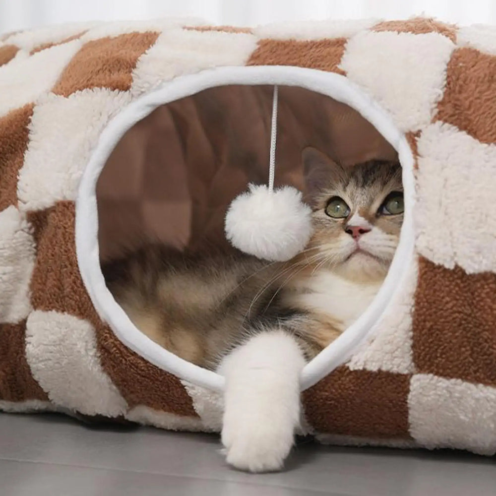 Brown and white checkerboard donut cat tunnel cave bed with hanging toy ball for indoor cats, styled under a decorated Christmas tree